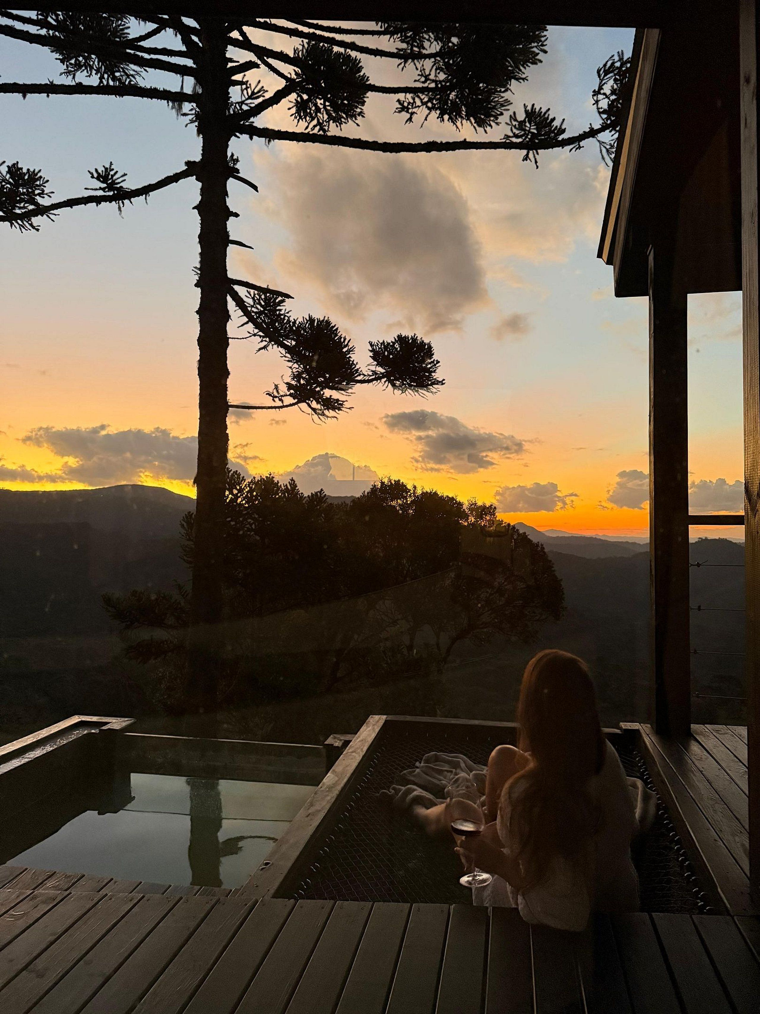 A person relaxing on a wooden deck with a glass of wine, overlooking a scenic sunset view with mountains and trees.