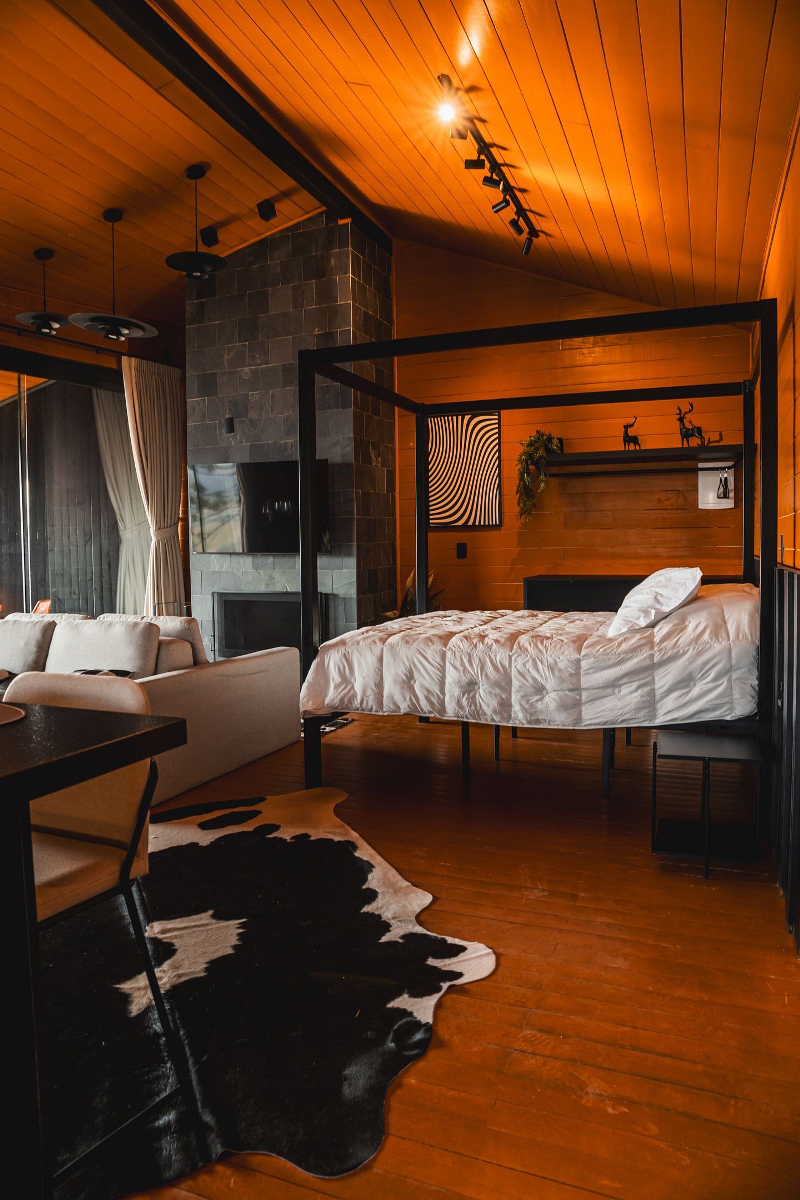 Une chambre moderne avec une cheminée en pierre, un lit recouvert de linge de maison blanc, un tapis en peau de vache et un plafond en bois.