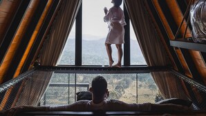 Ein Zimmer mit hoher Holzbalkendecke und einem großen Fenster. Eine Person steht am Fenster, während eine andere in einer Hängematte liegt.