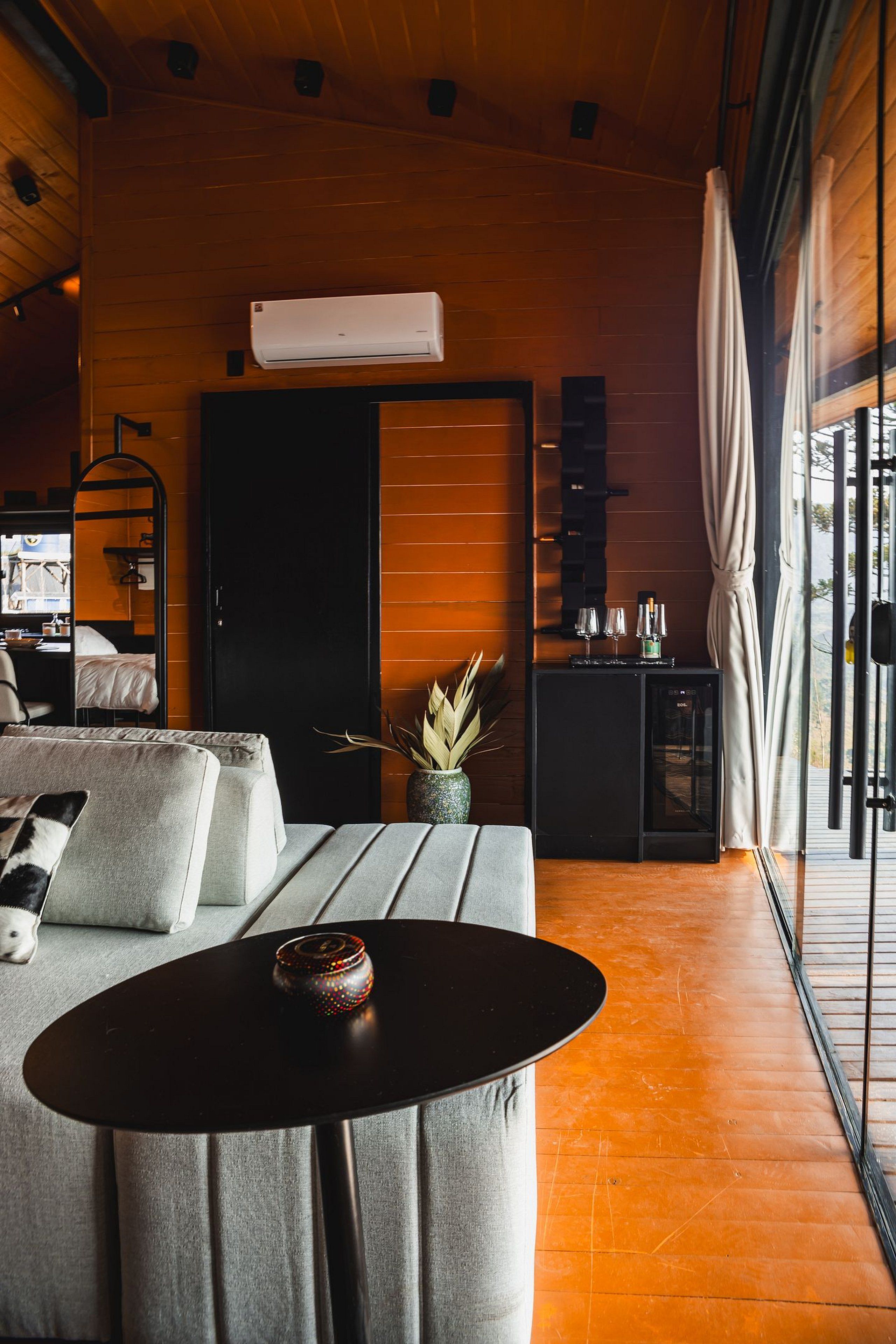 Una habitación de hotel moderna con techo de madera, un ventanal con cortinas, un sofá con cojines, una mesita con un objeto decorativo y un minibar.