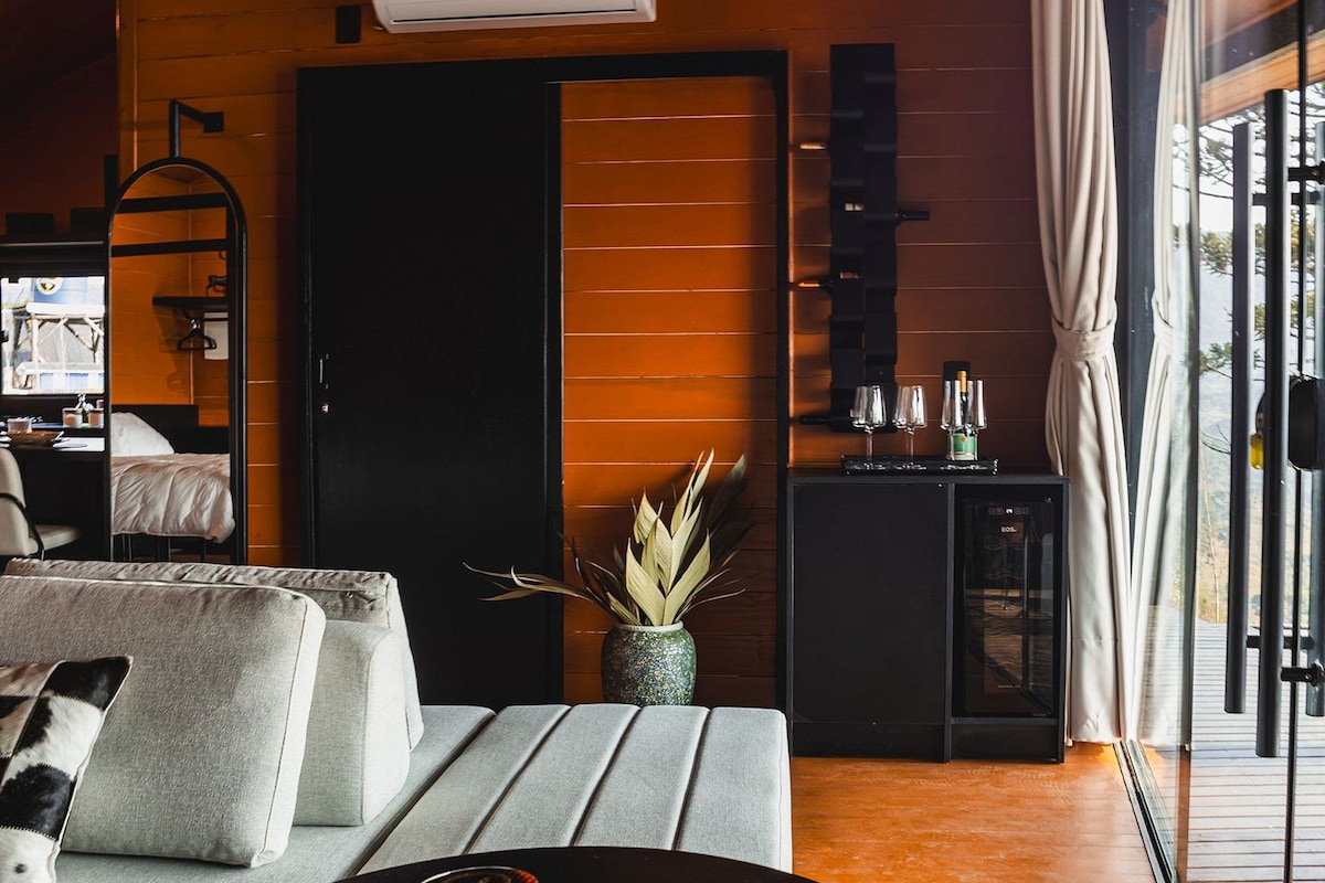 Ein modernes Hotelzimmer mit Holzboden, einem großen Fenster mit Vorhängen, einer Couch mit Kissen, einem kleinen Tisch mit Dekoration und einer Minibar.