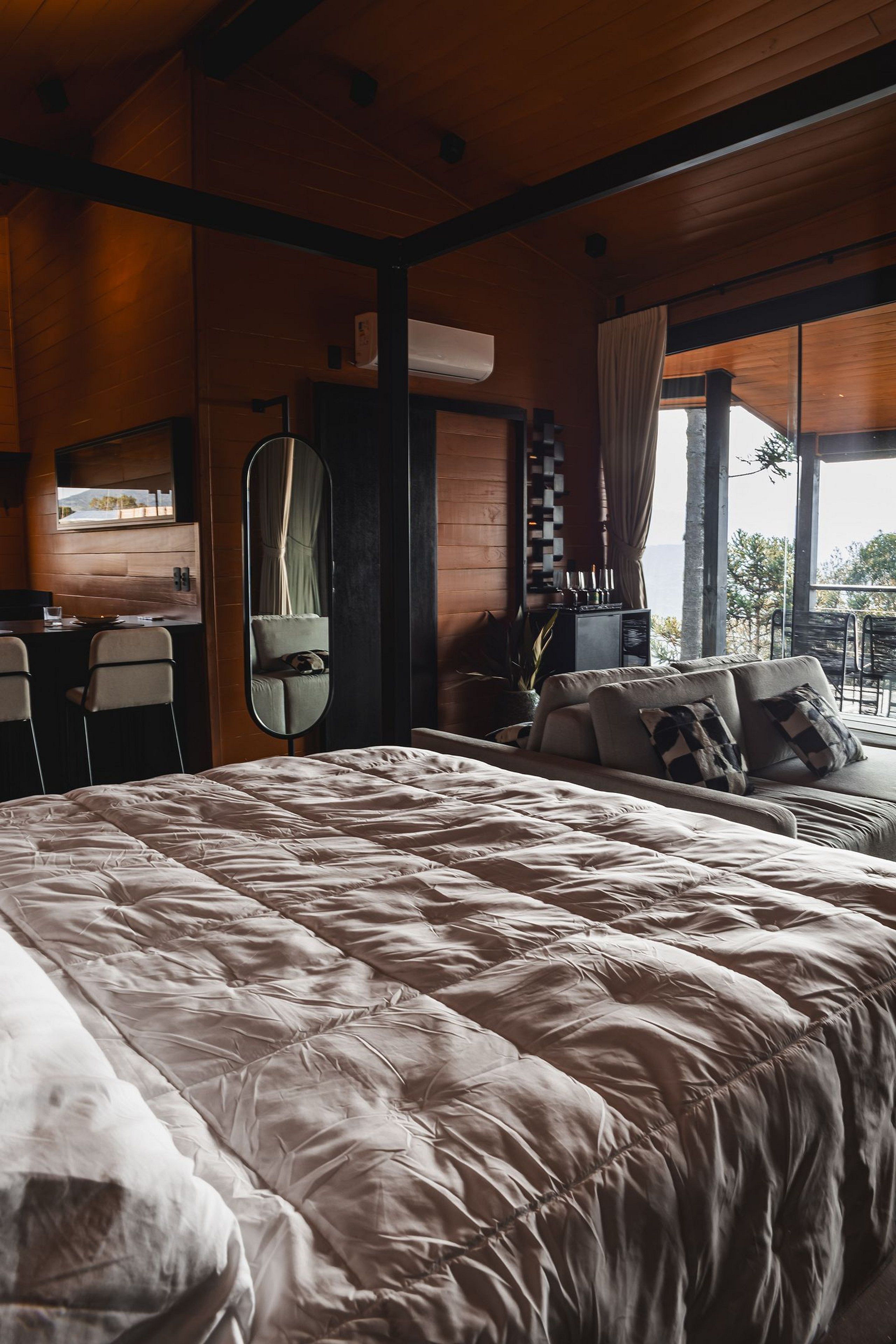 A bedroom with a large bed, a desk, a chair, a mirror, and a view of the outdoors.