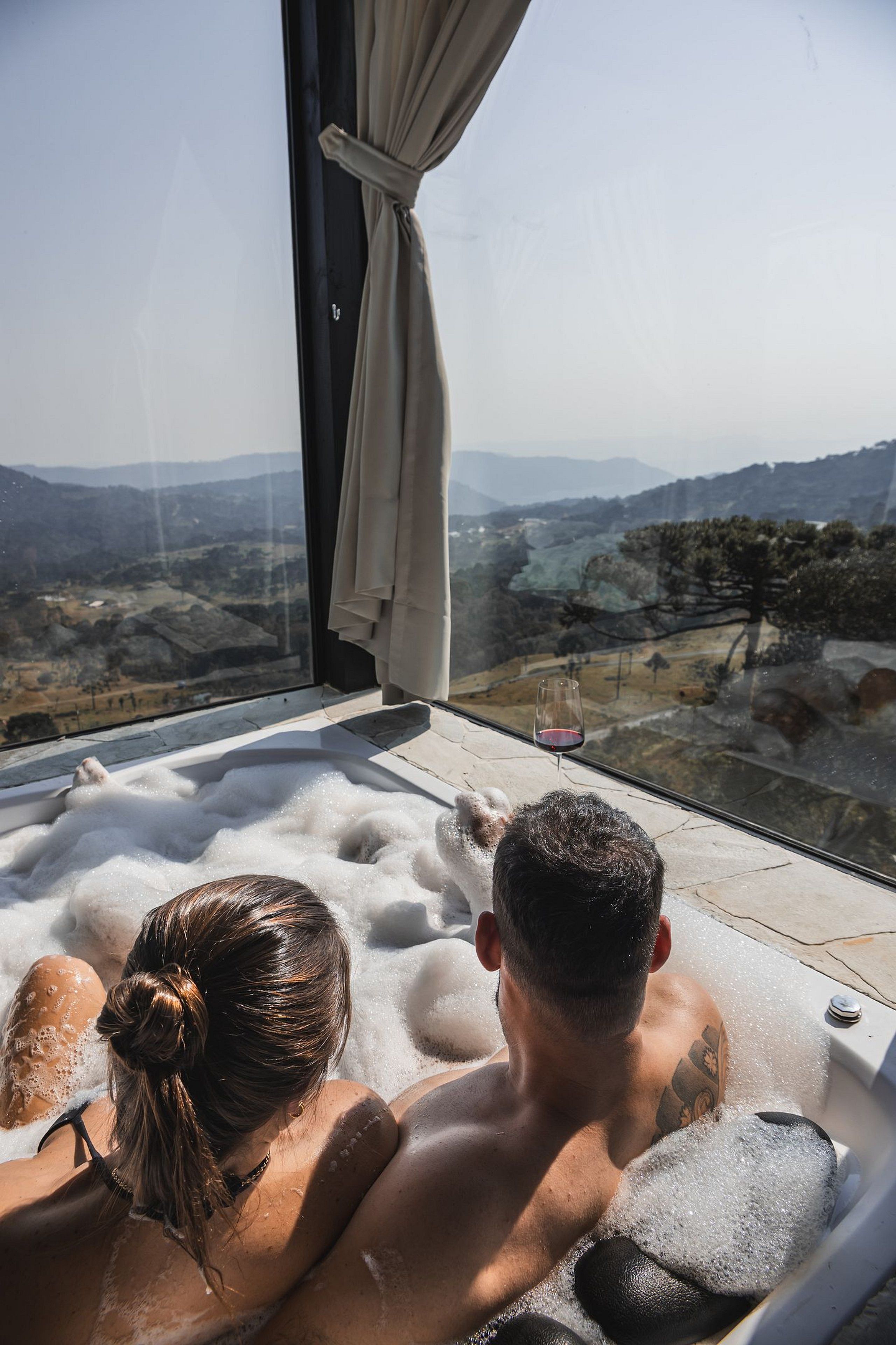 Um casal em banheira de hidromassagem com vista panorâmica de um vale e montanhas.