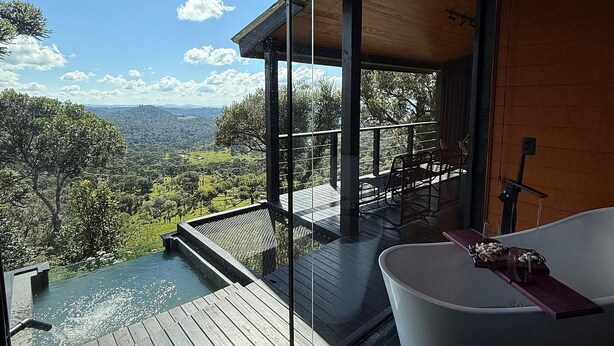 Ein modernes Badezimmer im Freien mit einer freistehenden Badewanne, einer Holzterrasse und einem gläsernen Bereich mit Blick auf eine reizvolle Landschaft.