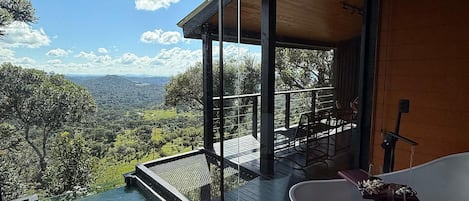 Een moderne, openlucht badkamer met een vrijstaand bad, een houten terras en een glazen ruimte met uitzicht op een prachtig landschap.