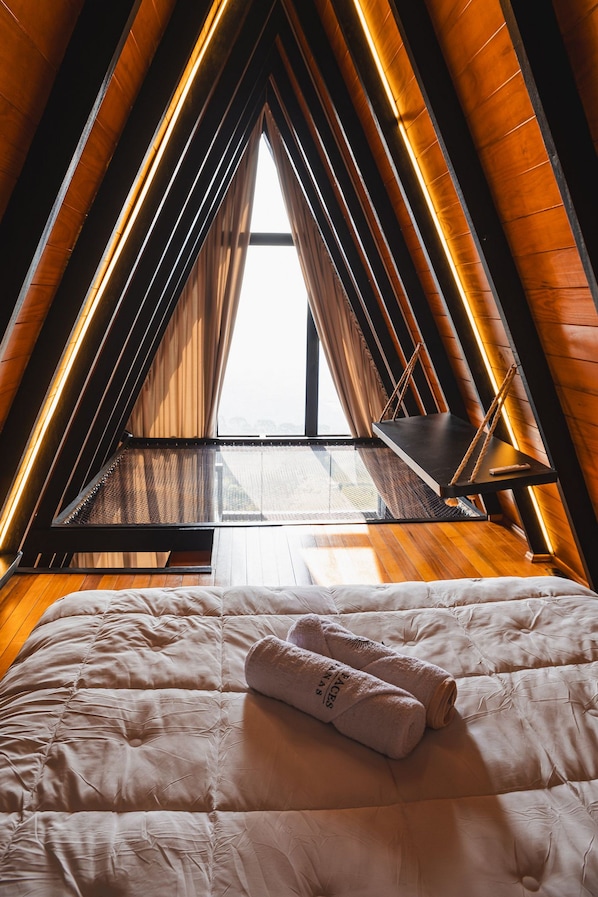 A bedroom with a sloped ceiling, a bed with a white duvet, and a window with a view of the city. - Cabana Duas Faces (Urubici)