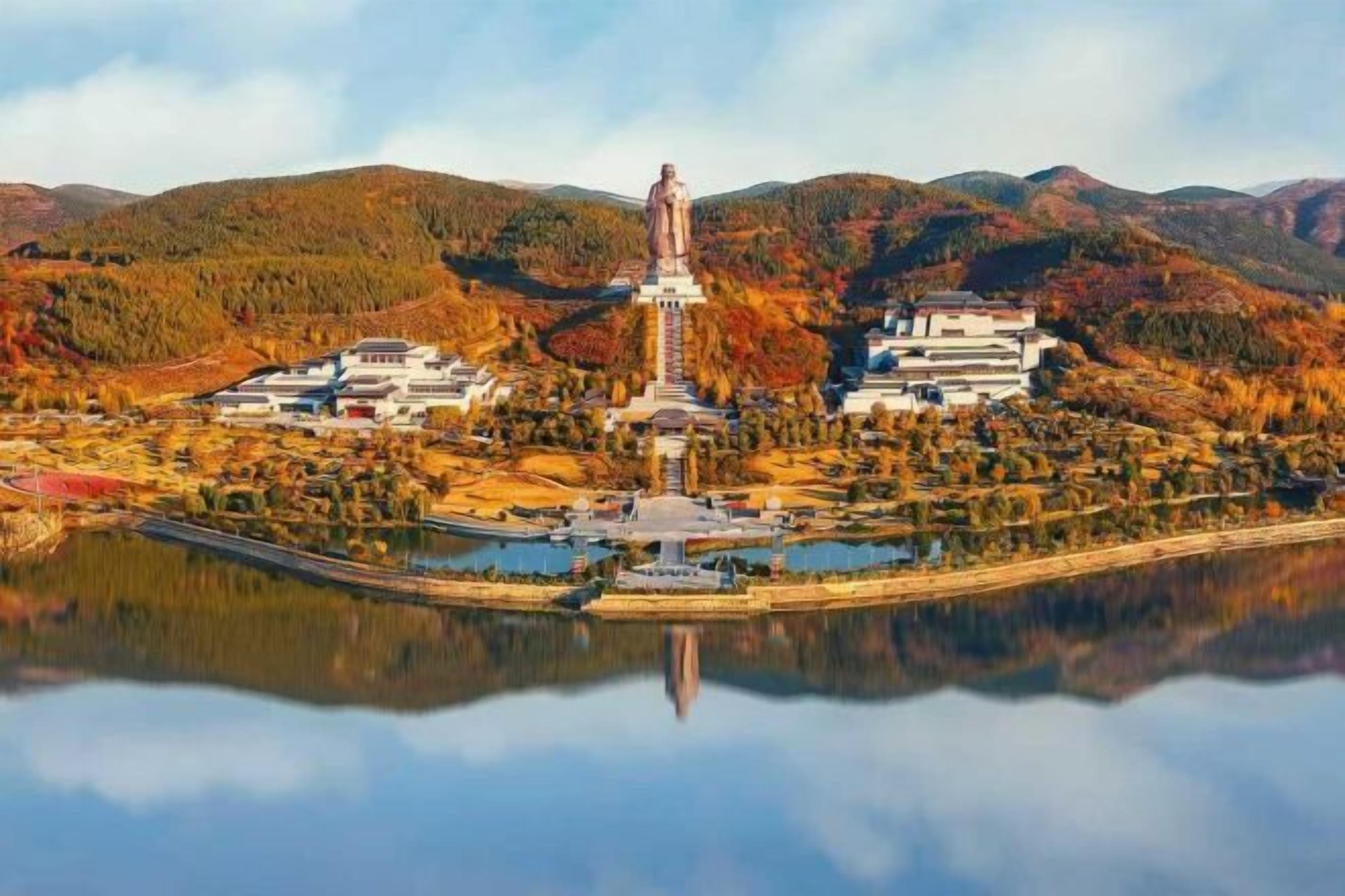 A large statue in a mountainous area with a reflection in a body of water, surrounded by buildings and trees.