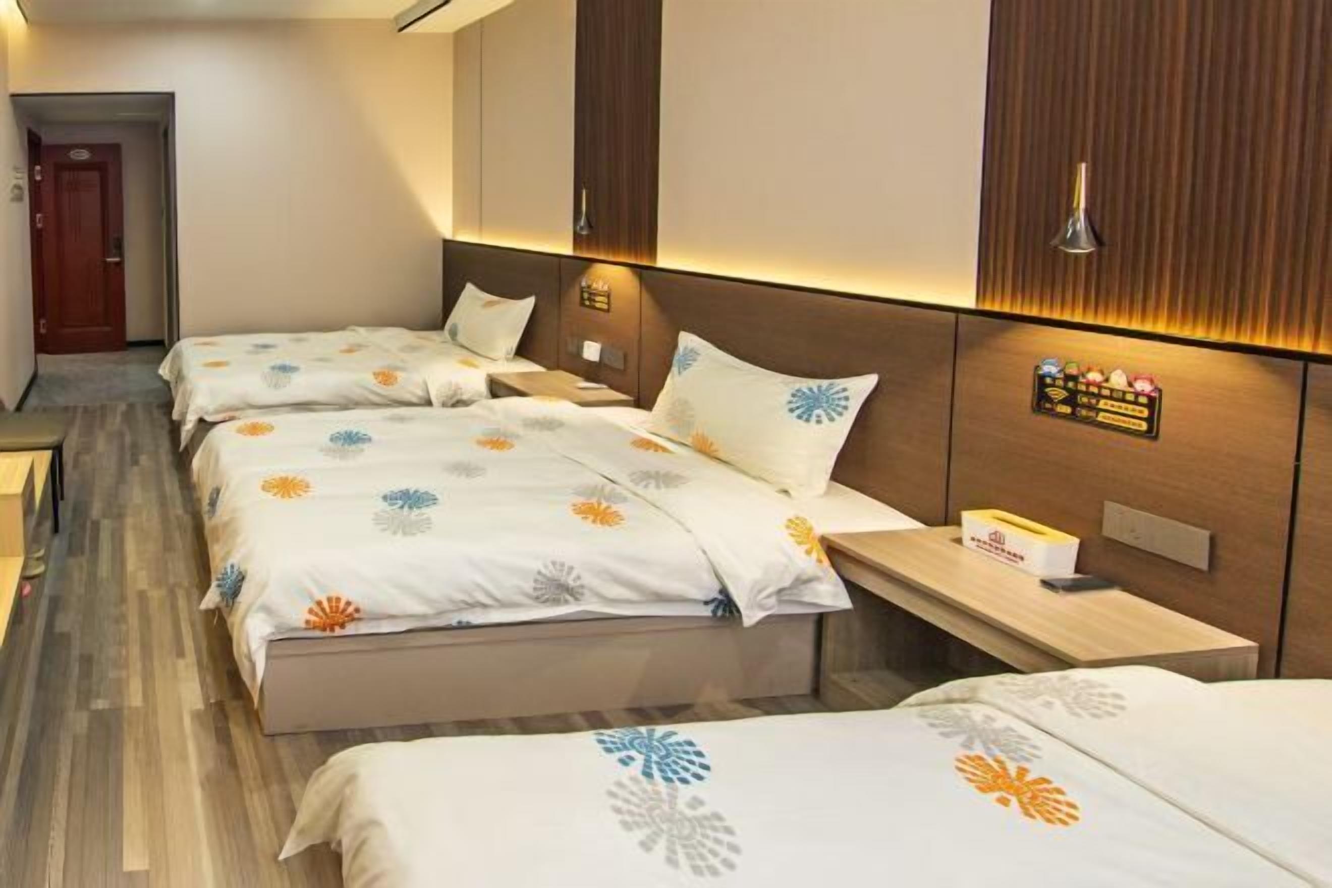 A hotel room with two beds, a wooden headboard, a bedside table with a lamp, and a small shelf with items.