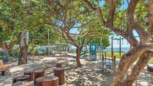 Un espace vert sablonneux agrémenté de troncs d’arbres, une aire de jeux avec un toboggan bleu et des arbres qui offrent de l’ombre.
