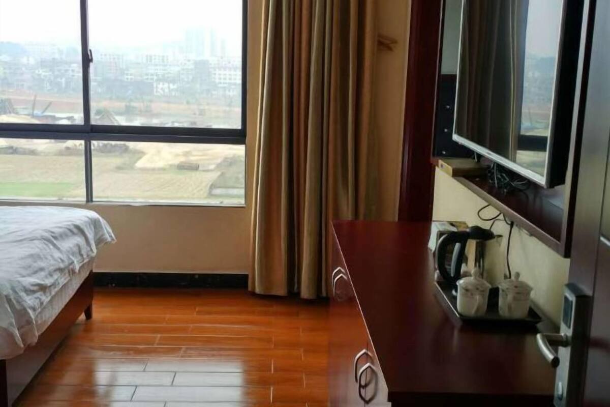 Quarto de hotel com cama, piso de madeira, televisão, cafeteira e janela com cortinas.