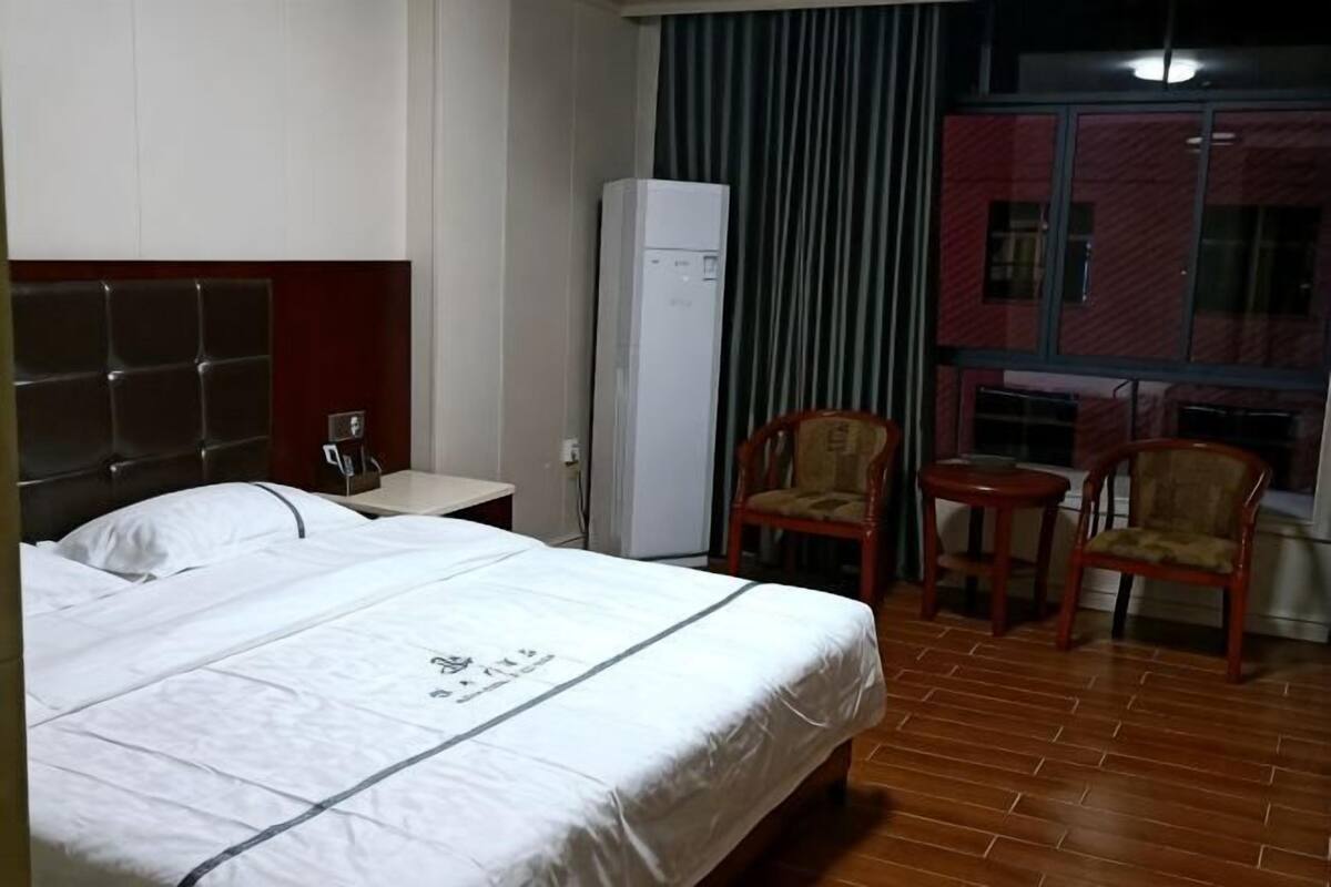 Quarto de hotel com cama, uma mesinha, duas cadeiras, frigorífico e janela com cortinas.