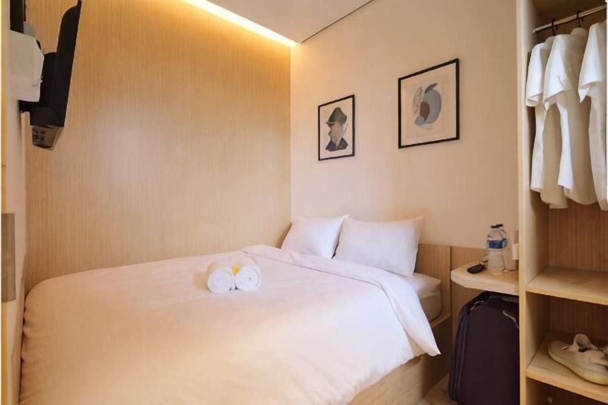 A hotel room with a bed, a wardrobe, a small table with a bottle, and two framed pictures on the wall.