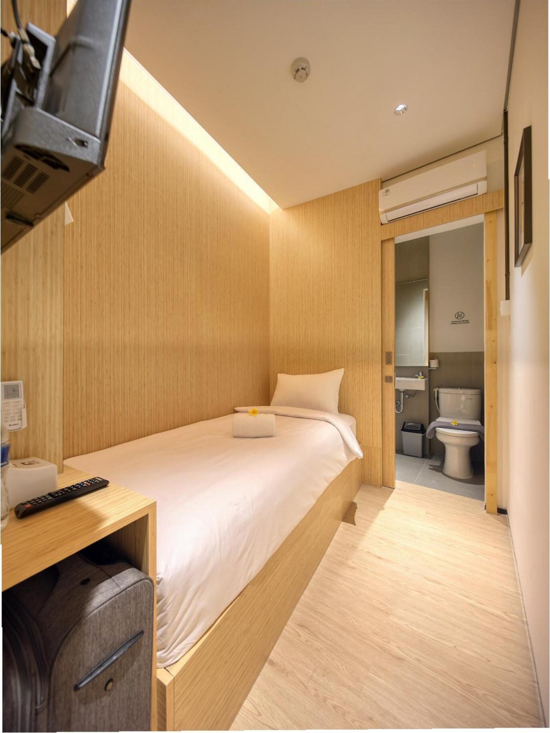 A compact hotel room with a bed, bedside table, and a small desk with a remote control.