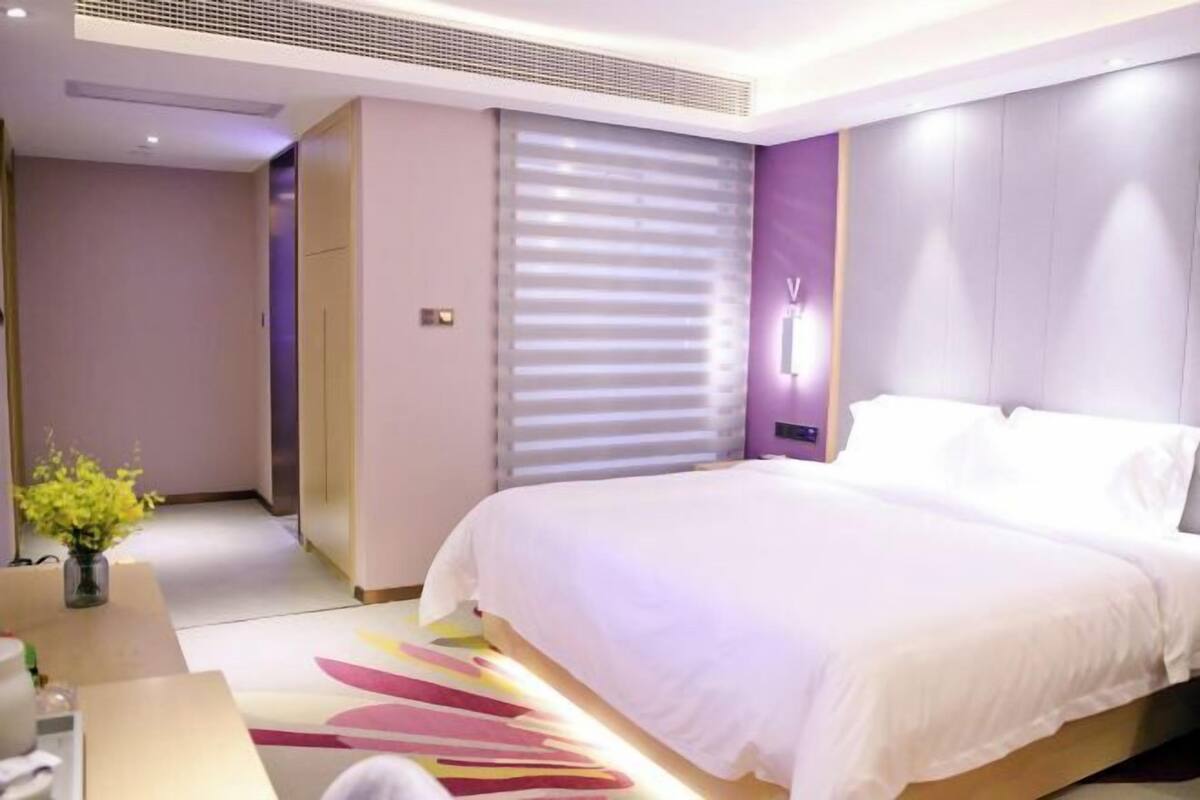 A modern hotel room with a large bed, a bedside table, a lamp, and a window with blinds.