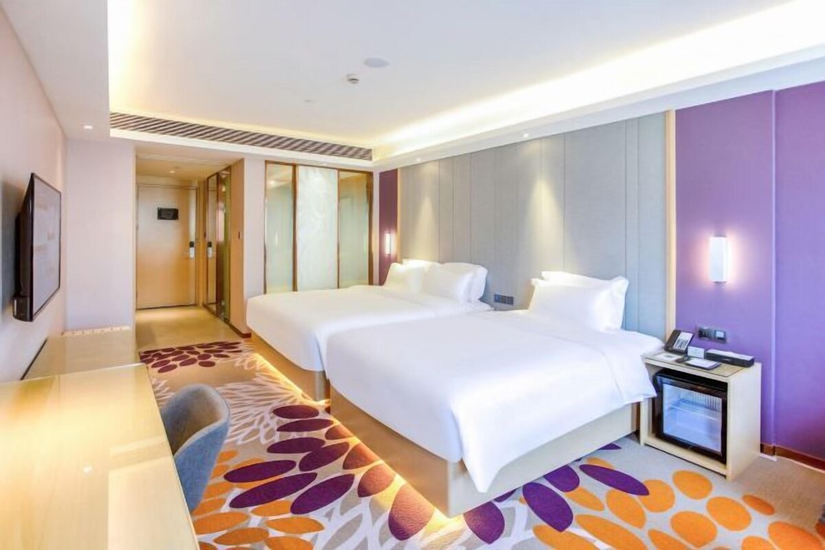 A hotel room with two beds, a desk, a chair, a small table with a lamp, and a television.