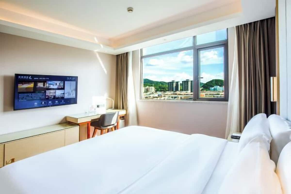 A hotel room with a bed, a desk, a chair, a TV mounted on the wall, and a large window offering a city view.