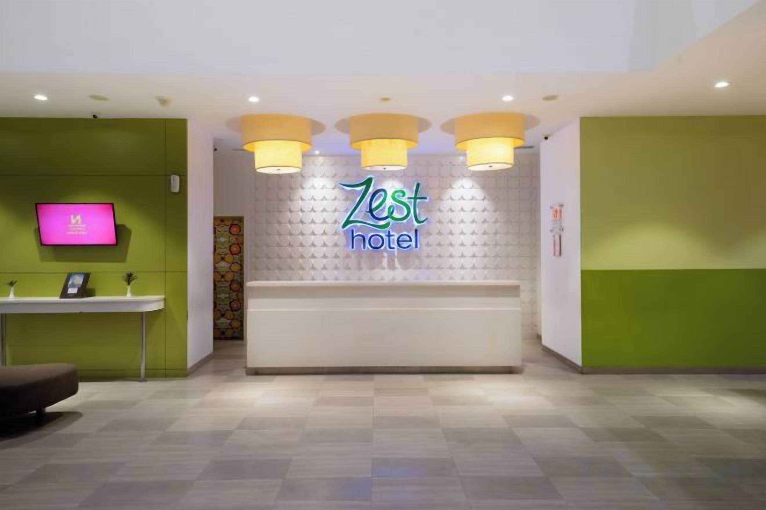 Un espace d’accueil d’hôtel comprenant un comptoir de réception, un téléviseur et un panneau indiquant « Zest Hotel ».