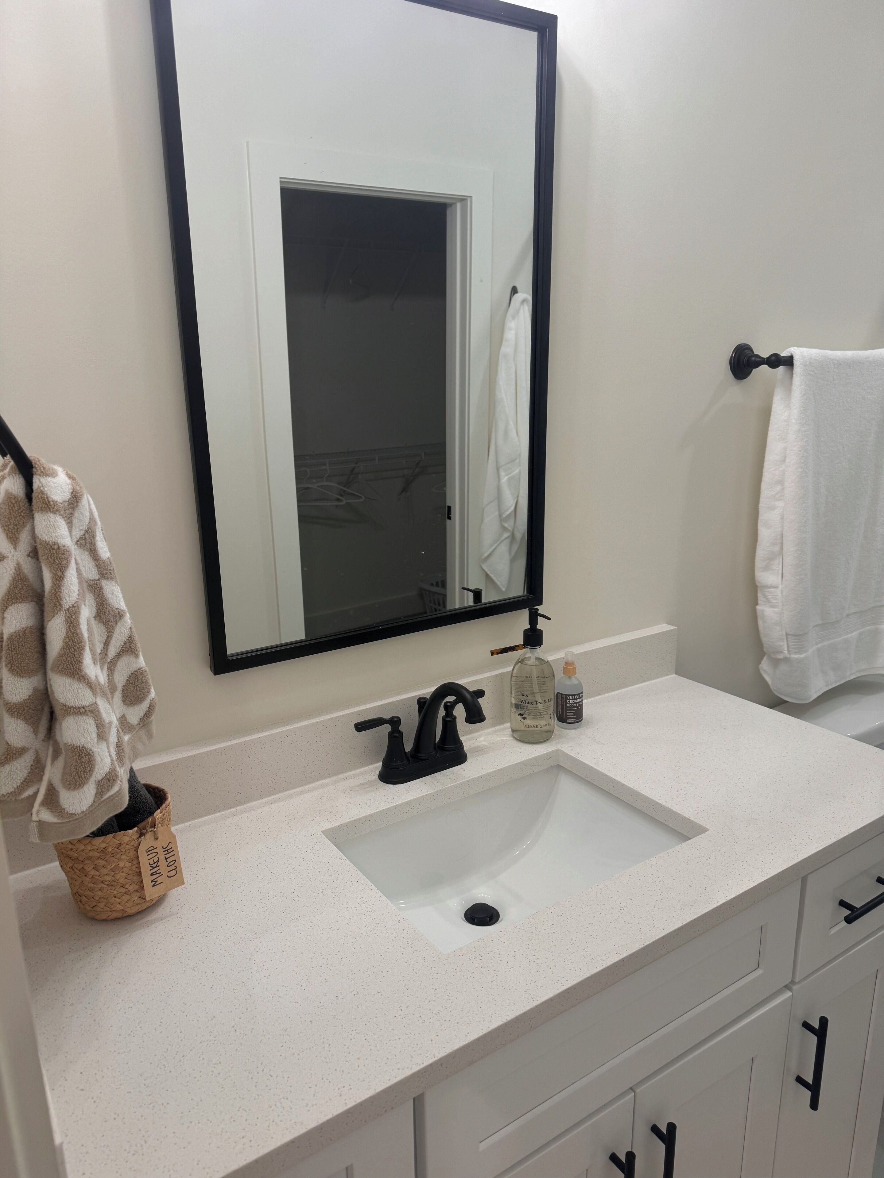 Une salle de bain avec un lavabo blanc, un robinet noir et un miroir au-dessus. On y trouve un porte-serviettes avec une serviette blanche et une serviette marron. Un flacon de savon liquide est posé sur le comptoir du lavabo.