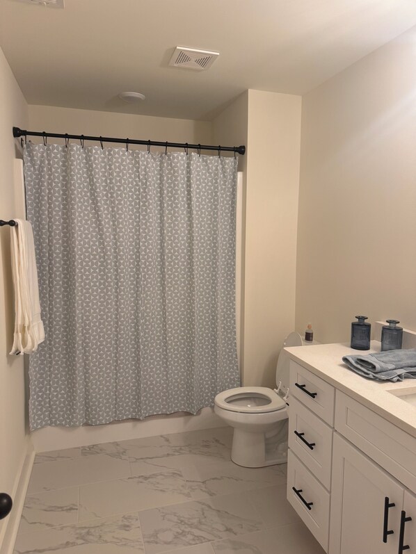 A bathroom with a toilet, a shower curtain, a marble-tiled floor, and a white vanity with drawers.