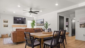 A modern living room with a brown sofa, wooden dining table, and chairs. There is a ceiling fan, a TV, and framed pictures on the walls. - Charming 3 Bedroom Retreat: Perfect for Families, Travelers and Professionals (Billings)