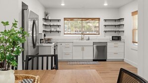 A modern kitchen with white cabinets, a stainless steel refrigerator, a microwave, and a sink. There is a window providing natural light and a plant on the left side. - Charming 3 Bedroom Retreat: Perfect for Families, Travelers and Professionals (Billings)