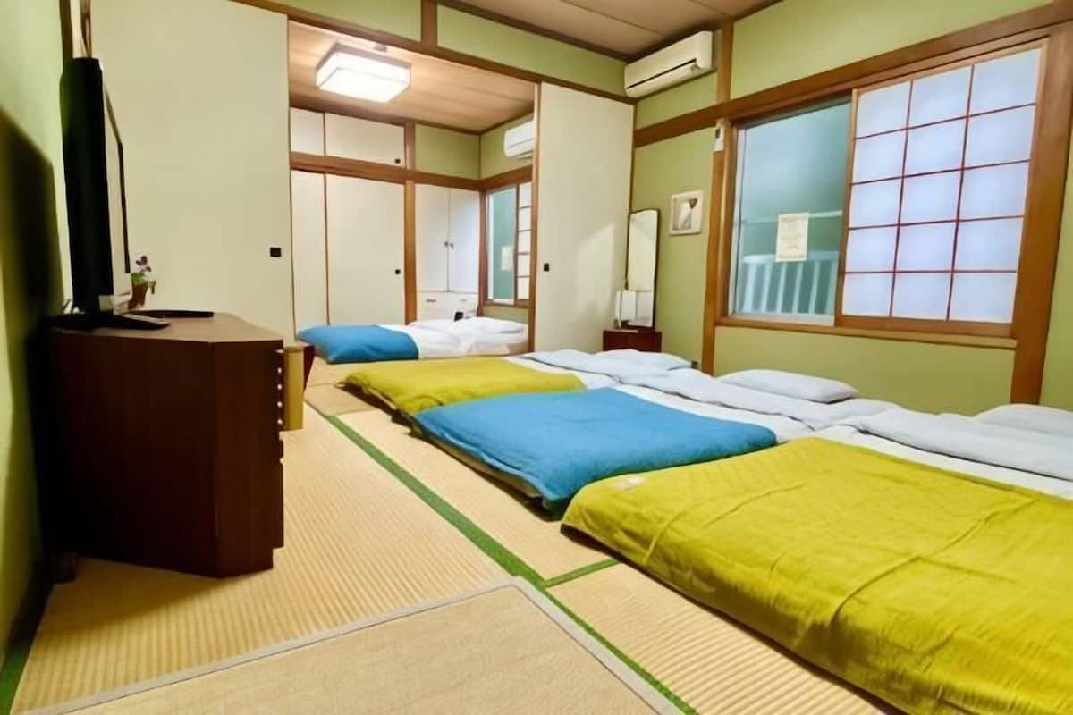 A room with tatami flooring, a desk, three beds with different colored bedding, a mirror, and a window with a grid pattern.