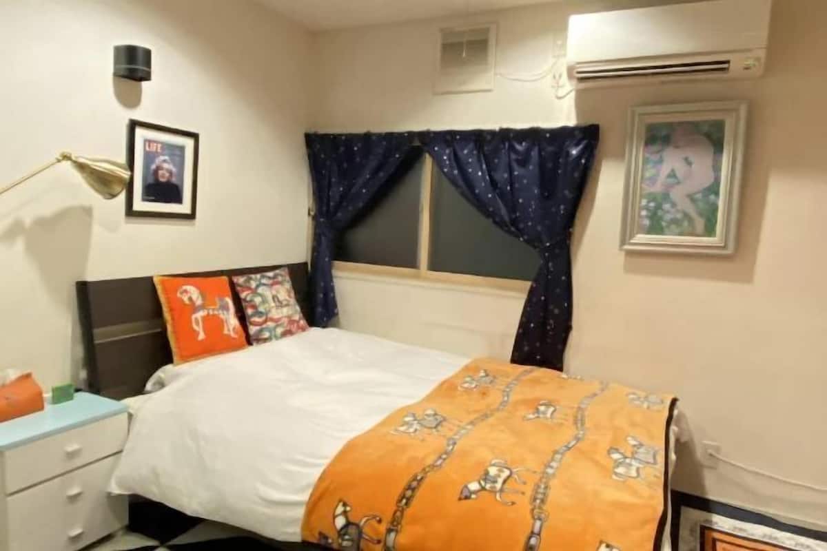 A bedroom with a bed, a nightstand, a lamp, a window with curtains, and a wall-mounted air conditioner.