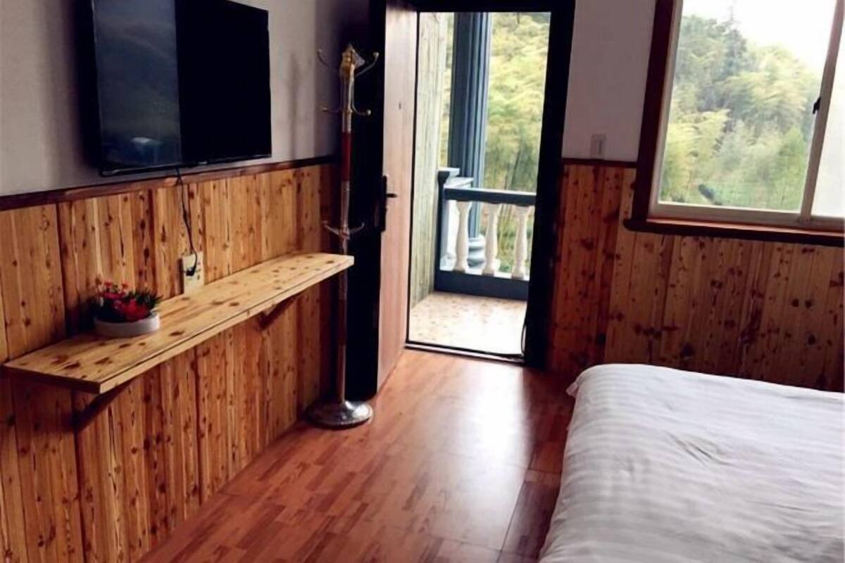 Une chambre avec un lit, un téléviseur fixé au mur, une étagère en bois avec un panier de fruits et une porte-fenêtre donnant sur un espace extérieur.