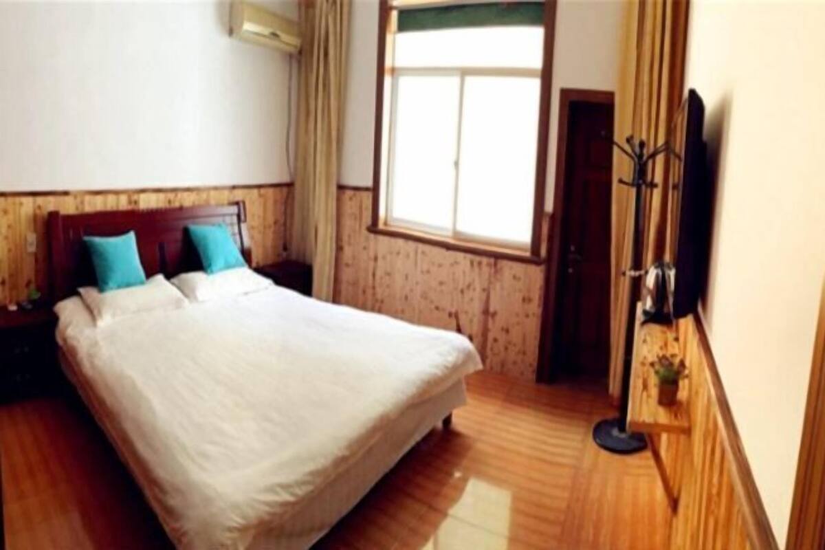 Une chambre d’hôtel avec un grand lit, un plancher en bois, une fenêtre avec des rideaux, un téléviseur à écran plat fixé au mur et une petite table avec une plante.