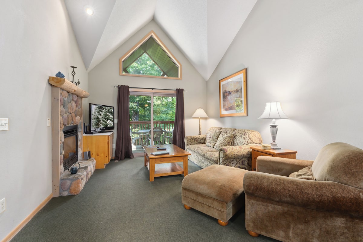 A cozy living room with a stone fireplace, a wooden coffee table, a patterned sofa, and a large window with curtains.