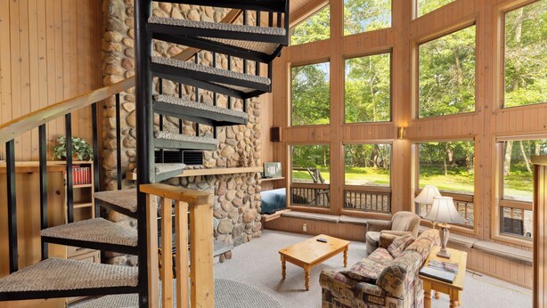 A cozy interior with a stone fireplace, wooden staircase, large windows, and comfortable seating. - Causeway on Gull (Nisswa)