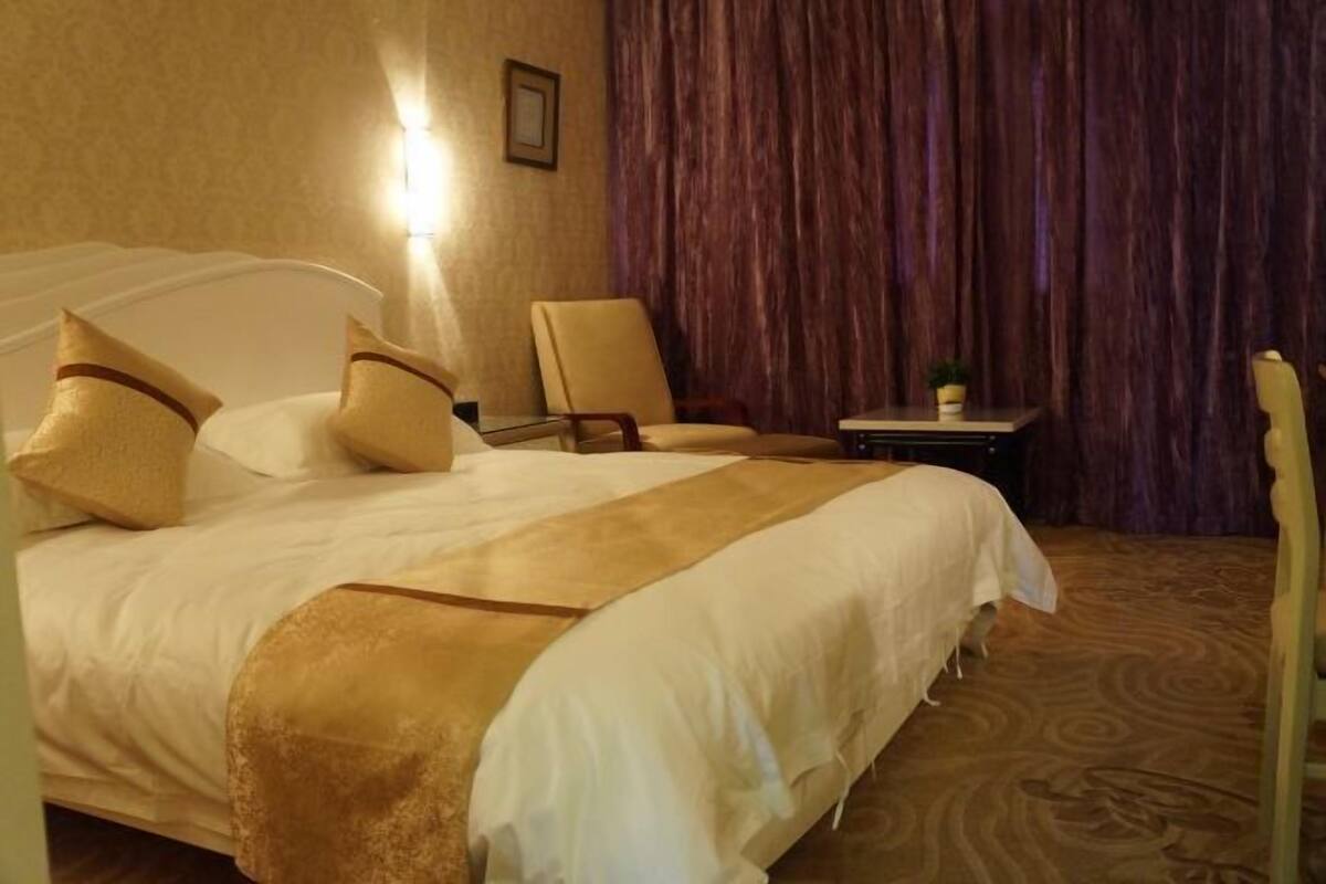 Quarto de hotel com uma cama grande, uma cadeira, uma mesinha e uma cortina.