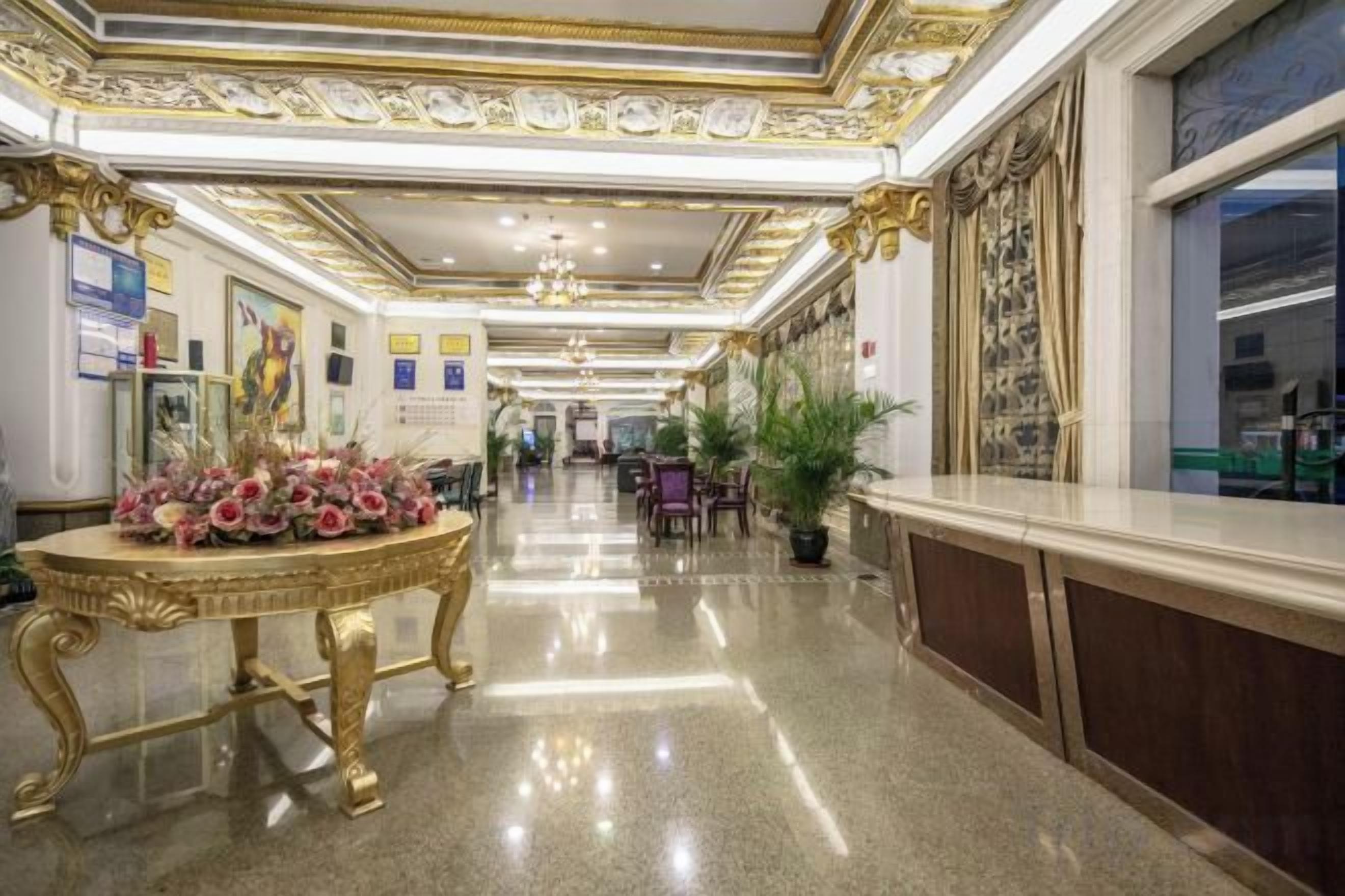 A hotel corridor with a polished floor, a gold-framed table with flowers, and a view of the lobby in the distance.