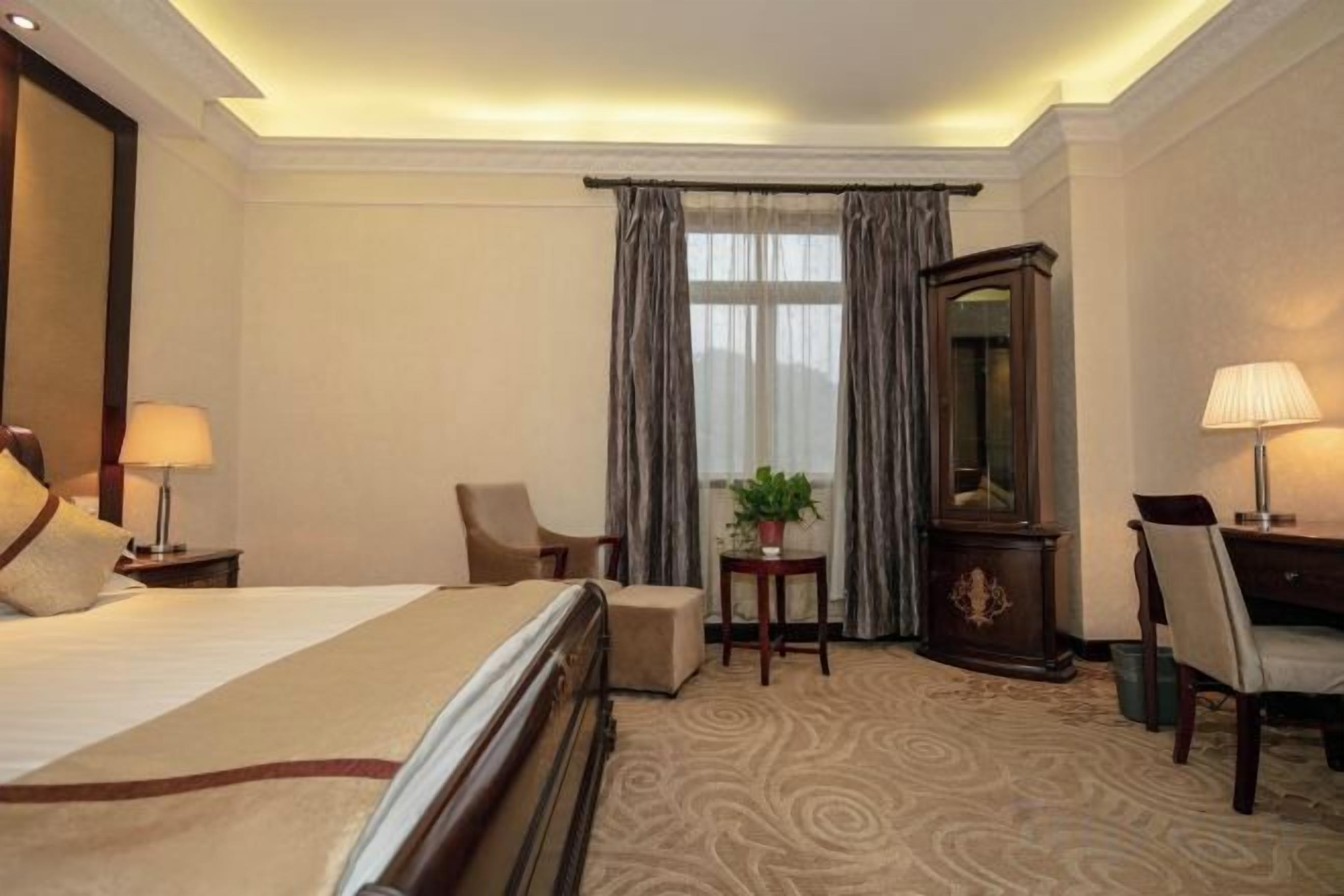 A hotel room with a bed, two chairs, a desk, a lamp, a mirror, and a window with curtains.