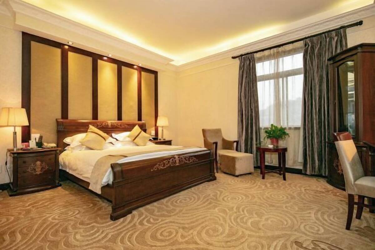 Quarto de hotel com uma cama grande, uma mesa de cabeceira, uma cadeira, uma pequena mesa com uma planta e vista para o exterior pela janela.