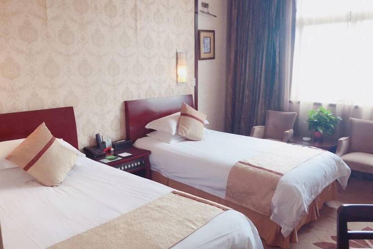 Quarto de hotel com duas camas, mesa de cabeceira, cadeira, pequena mesa e janela com cortinas.