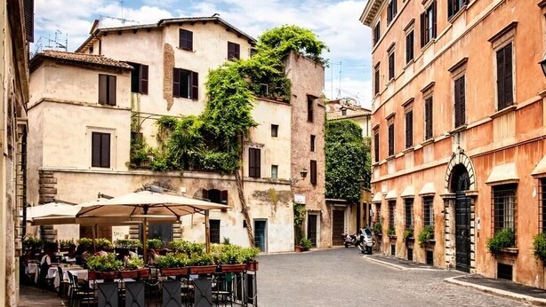 Une rue pavée bordée de bâtiments des deux côtés, des tables et chaises installées à l’extérieur sous des parasols, et un ciel dégagé.