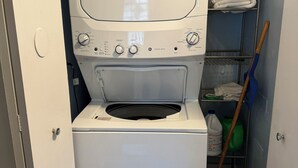 A compact washer and dryer unit in a laundry room with a metal shelf above containing folded towels.