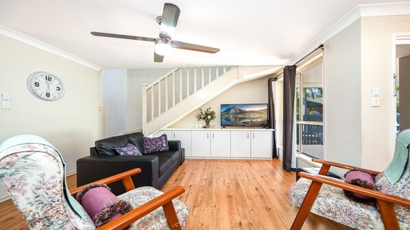 A living room with a black sofa, floral armchairs, a flat-screen TV, a ceiling fan, and a staircase.