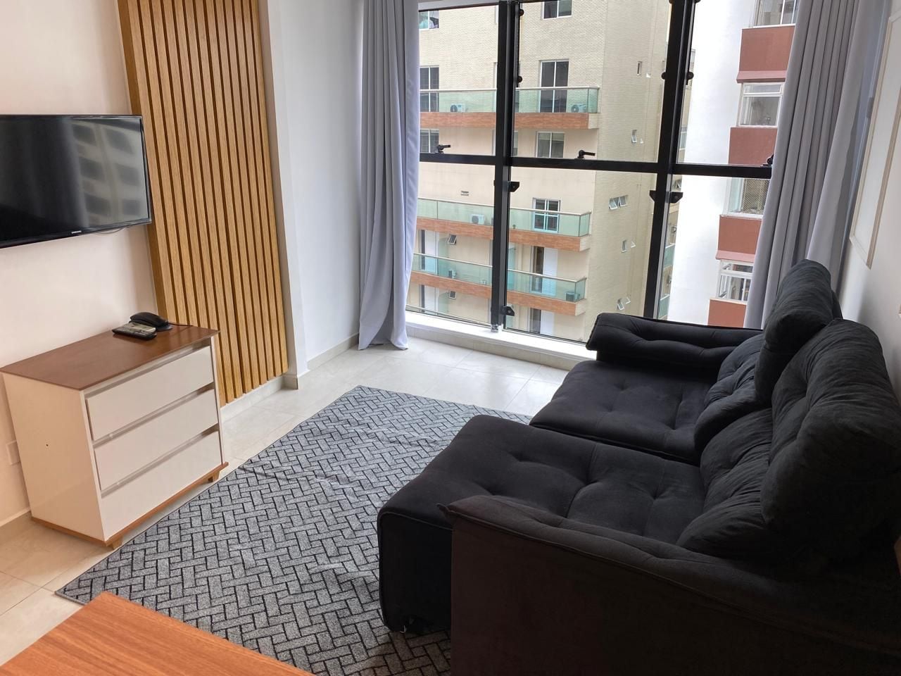 Una sala moderna con sofá, televisor en la pared, una mesita de madera y vista a un edificio a través de un ventanal grande.