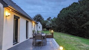 A modern house with a patio area featuring a table and chairs, surrounded by a well-maintained lawn and dense trees.
