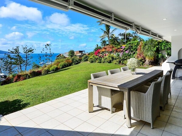 A patio with a dining table and chairs, a view of the ocean, and a well-maintained garden.