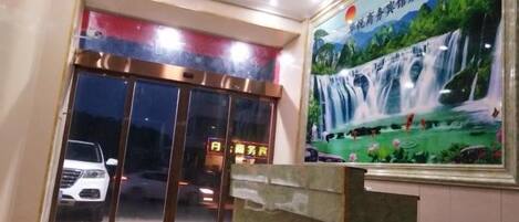 A hotel entrance with a glass door, a reception desk, and a waterfall painting on the wall.