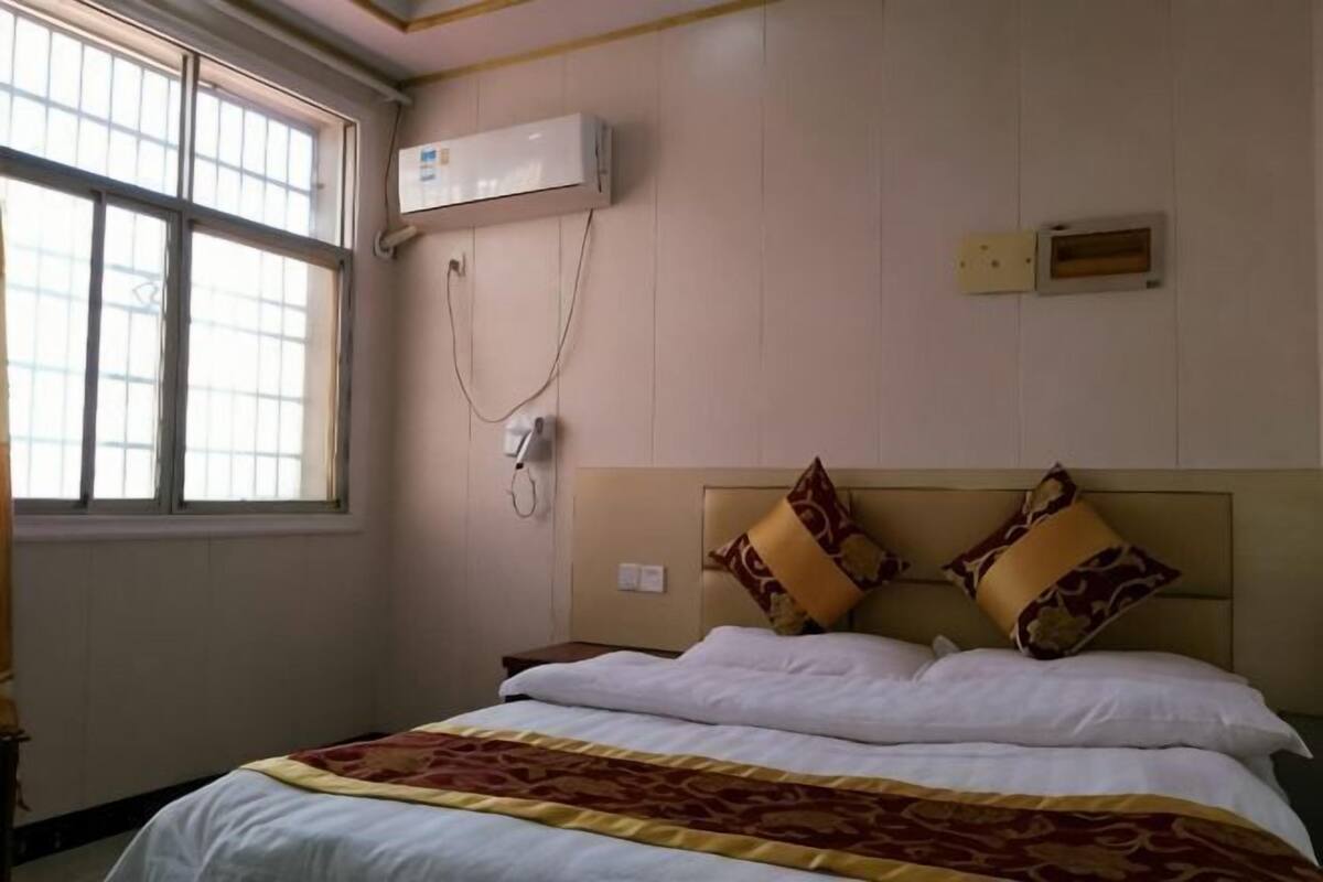 Una habitación de hotel con cama, ventana, aire acondicionado y un enchufe en la pared.