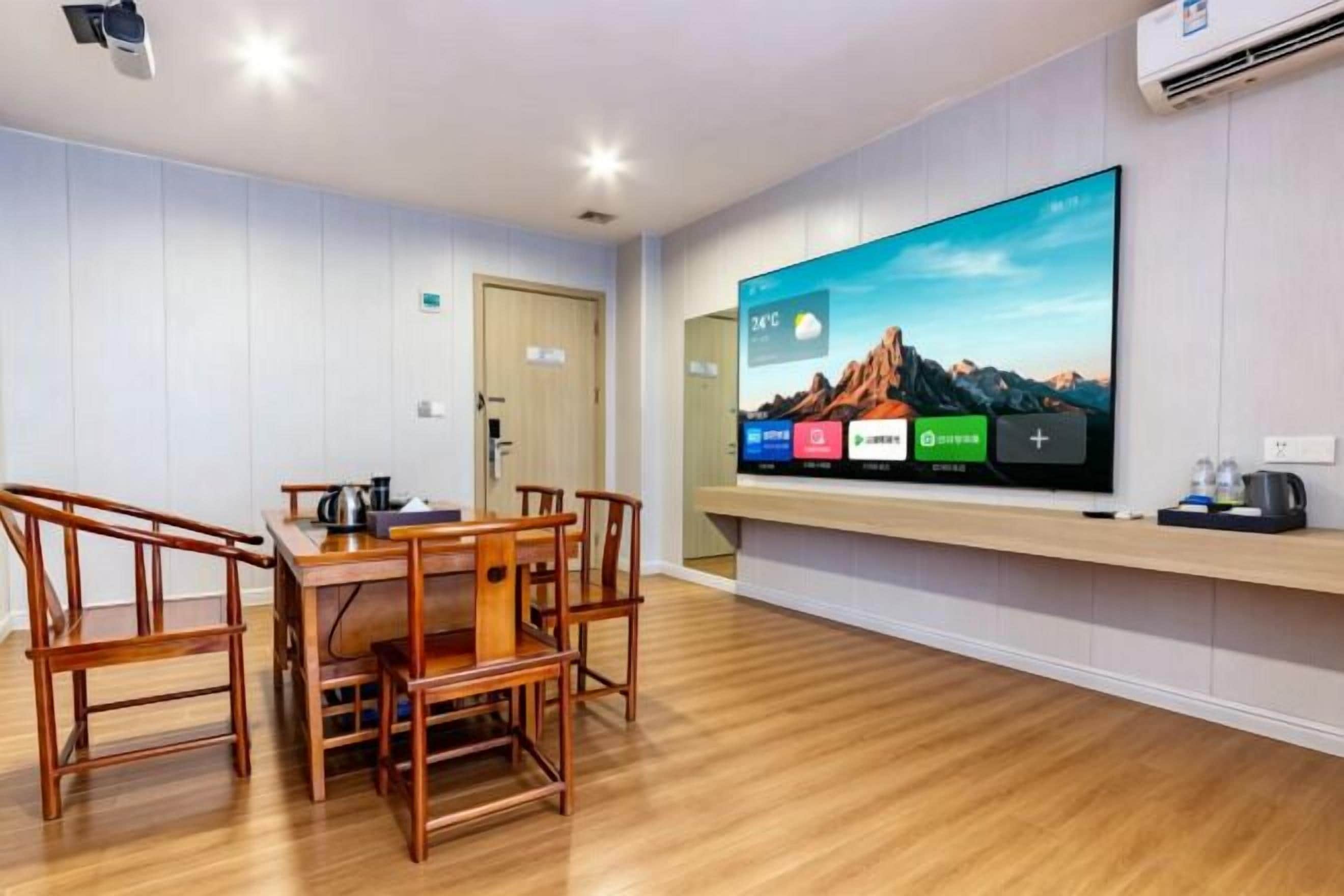 A hotel room with a wooden dining table, chairs, a flat-screen TV mounted on the wall, and a wooden floor.