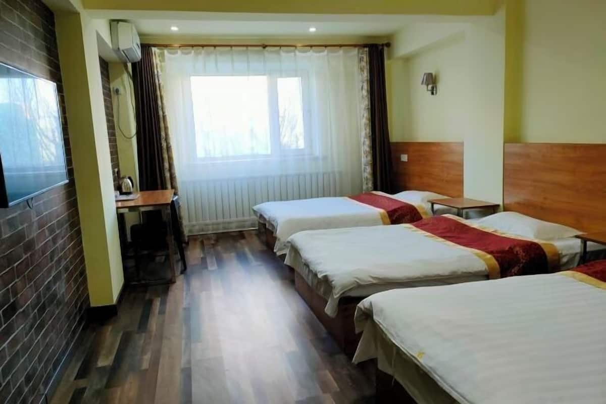 Ein Hotelzimmer mit drei Betten, einem Schreibtisch, einem Fernseher und einem Fenster mit Vorhängen.