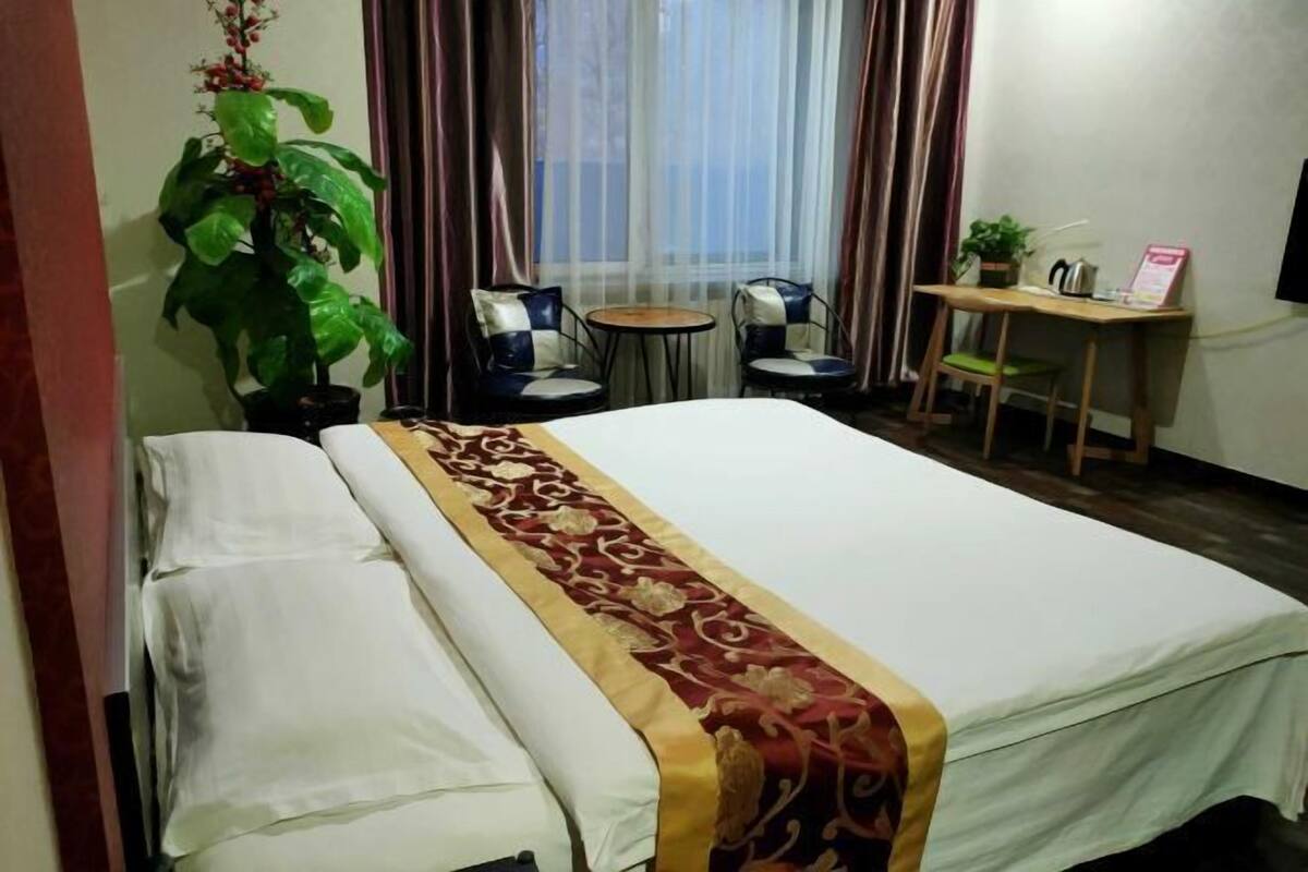 Ein Hotelzimmer mit einem Bett, einem Schreibtisch, zwei Stühlen, einem kleinen Tisch, einer Zimmerpflanze und einem Fenster mit Vorhängen.