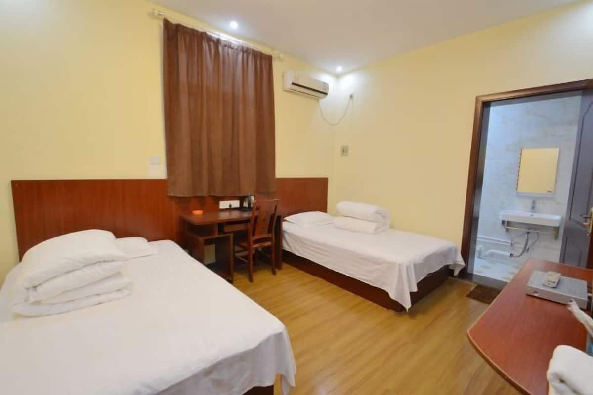 A hotel room with two beds, a desk, a chair, a wooden floor, an air conditioning unit, and a bathroom with a sink and mirror.