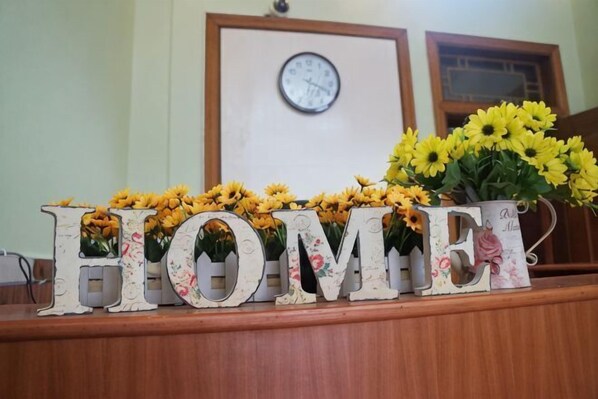 Un comptoir d’accueil avec le mot « HOME » épelé en lettres décoratives, une horloge au mur et un vase rempli de fleurs jaunes.