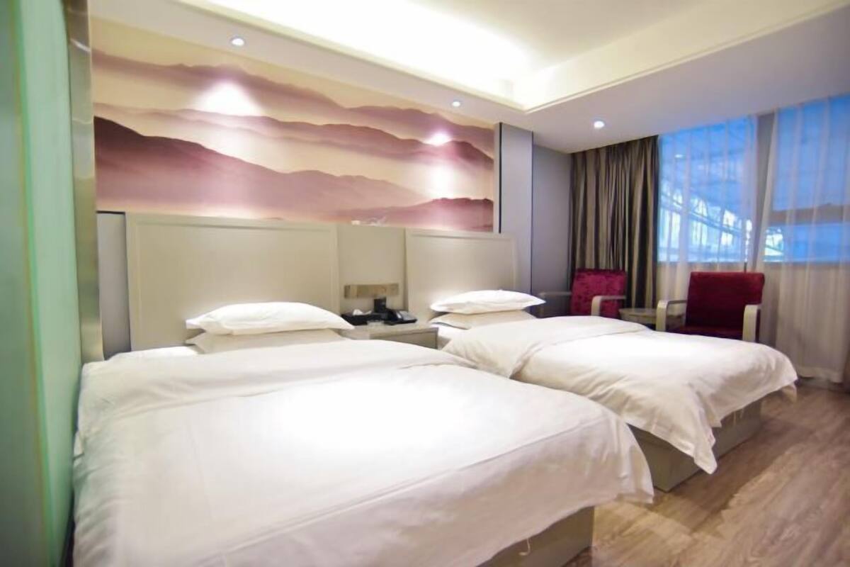 A hotel room with two beds, a large window with curtains, a red chair, and a wall with a painted landscape.